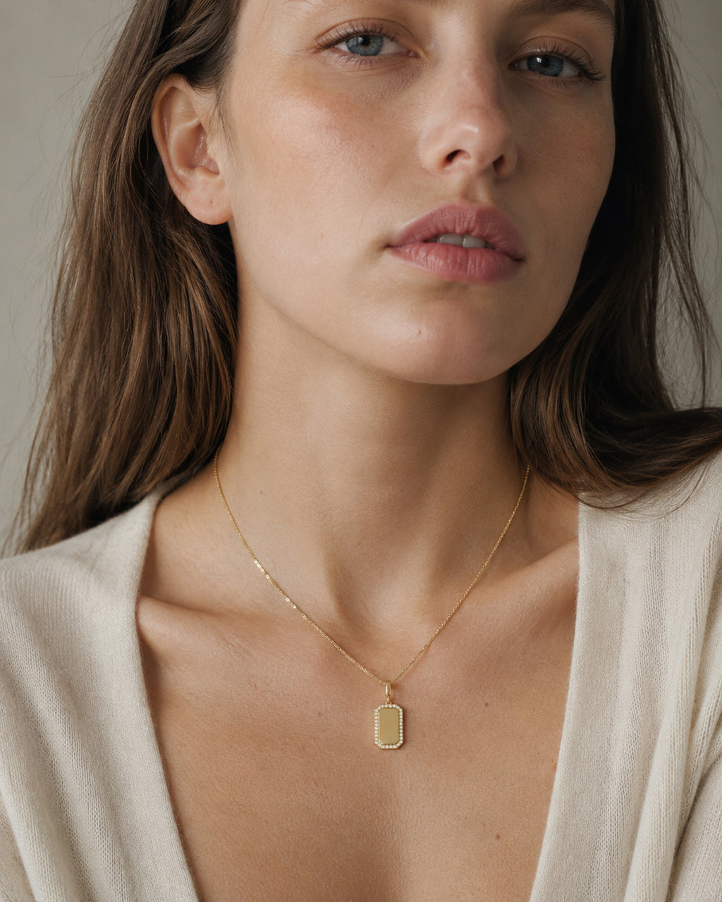 A close-up of a woman wearing a delicate gold chain necklace with a small rectangular gold pendant, styled with a cream-colored knit top against a neutral background.