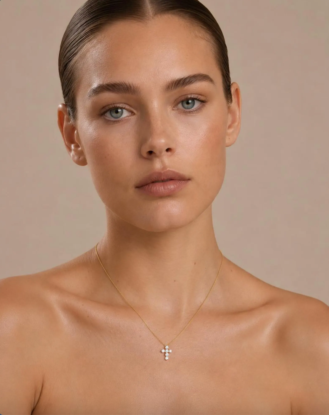 A close-up portrait of a woman with slicked-back hair wearing a delicate gold cross pendant necklace, set against a solid warm beige background.