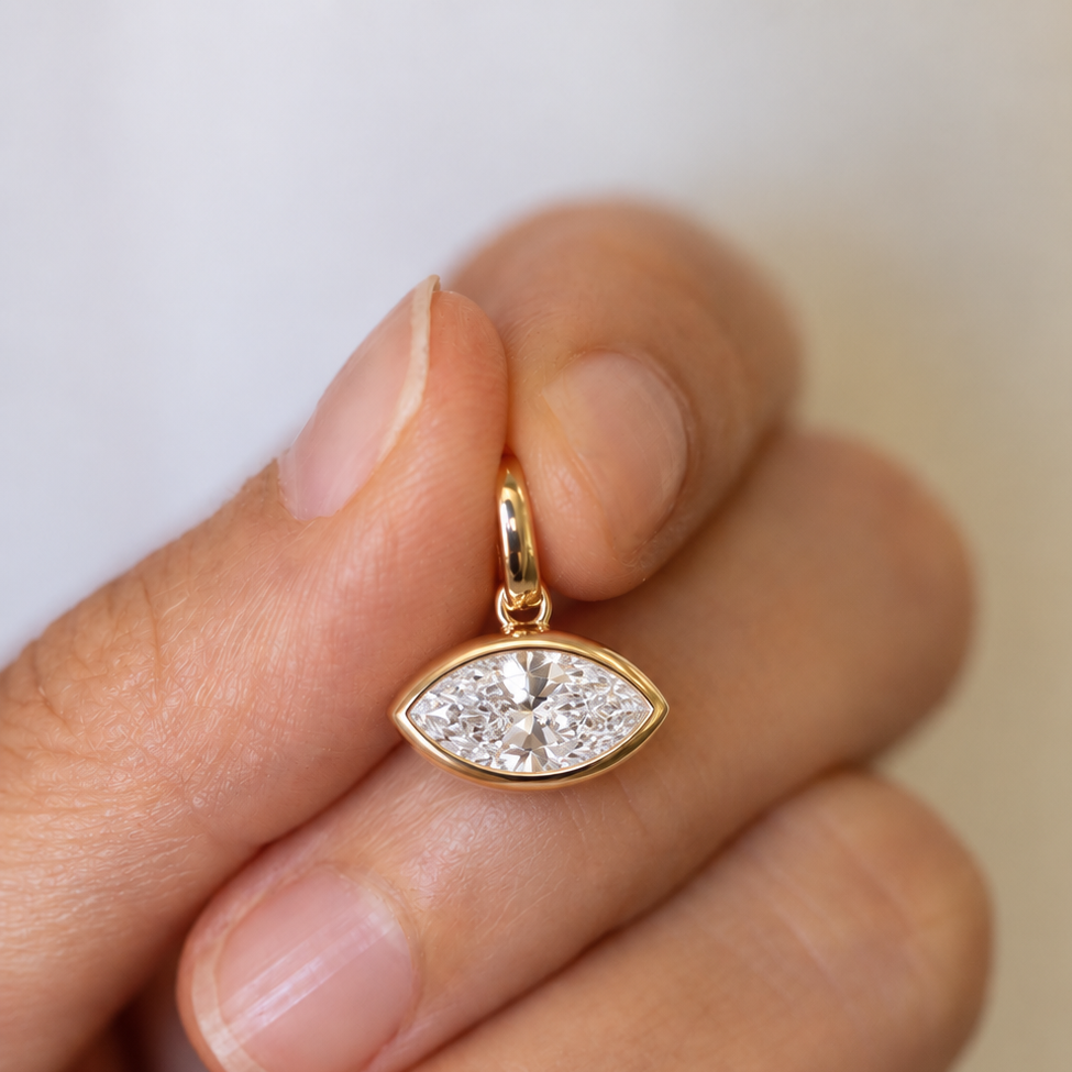 A close-up of a hand holding a small gold eye-shaped pendant with a clear marquise-cut gemstone, set against a soft, out-of-focus light background.